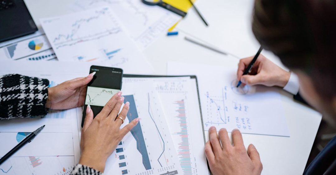 Pandangan dari atas orang-orang yang menganalisis grafik dan data keuangan dengan ponsel pintar dan dokumen.